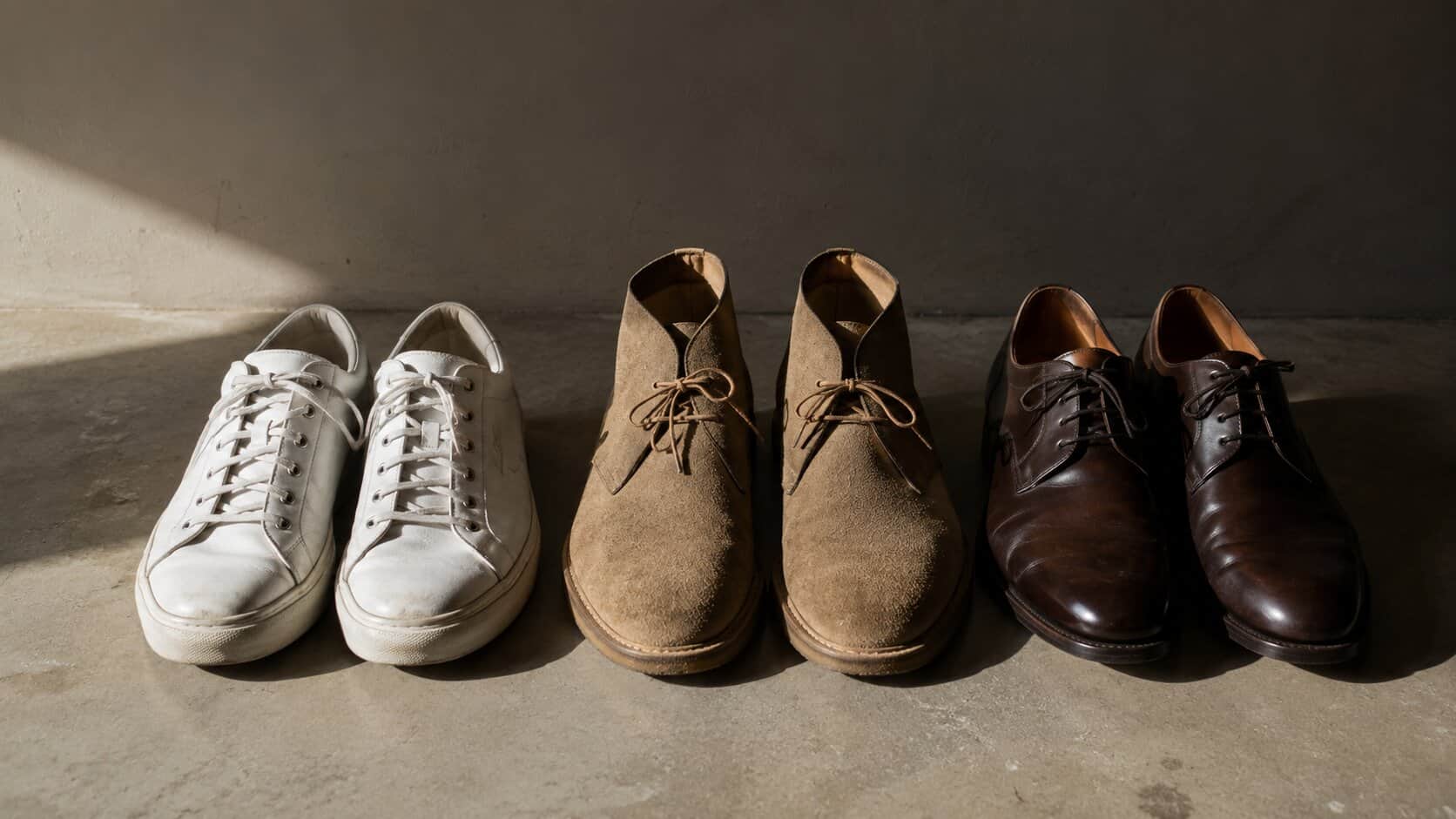 Three pairs of men’s shoes side by side on a bare floor, lit from one side: white leather sneakers, tan suede desert boots, and polished dark brown derbies.