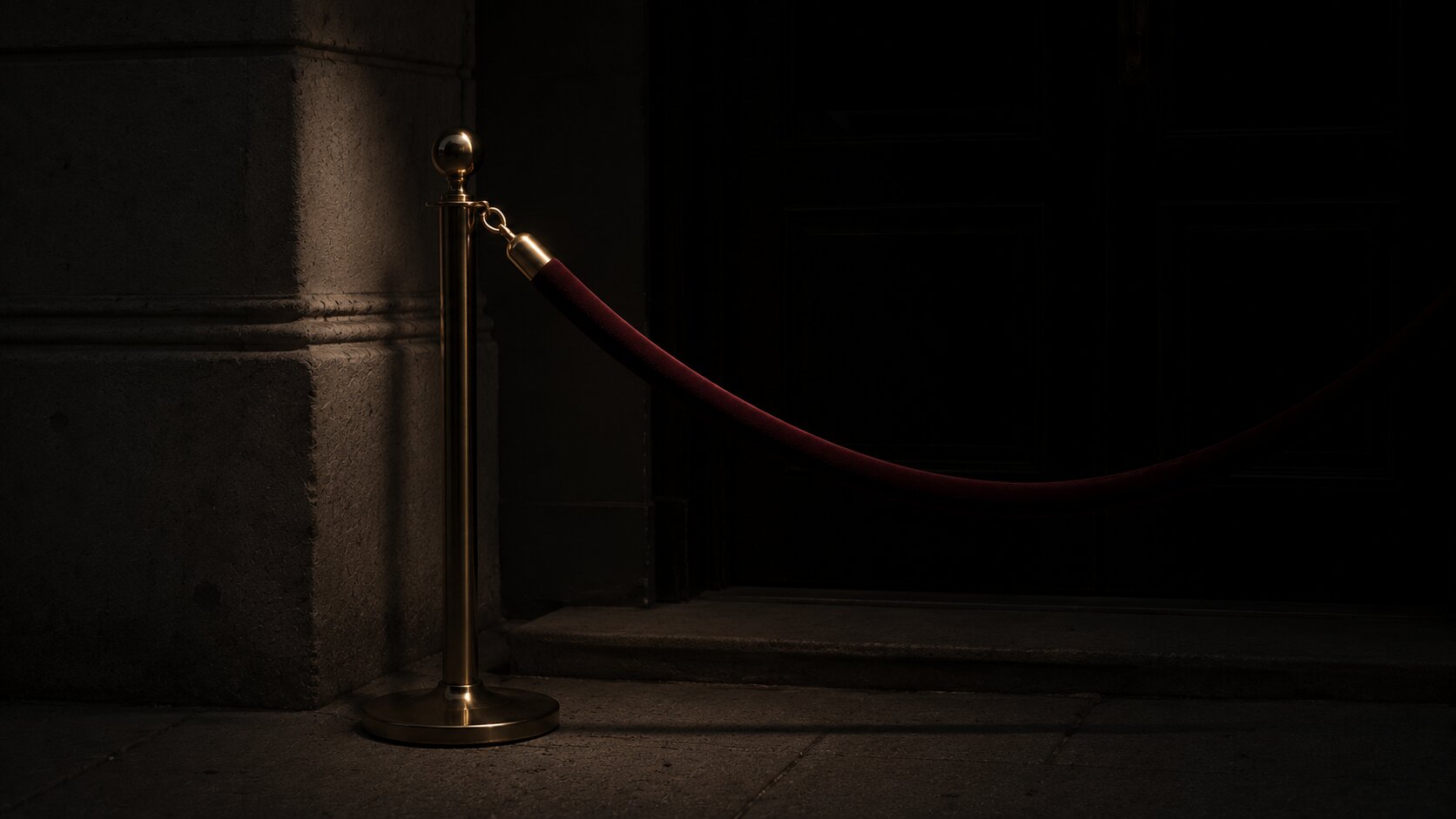 A velvet rope and a stanchion outside a dark doorway, a single overhead light picking out the brass clip where the rope hooks on.
