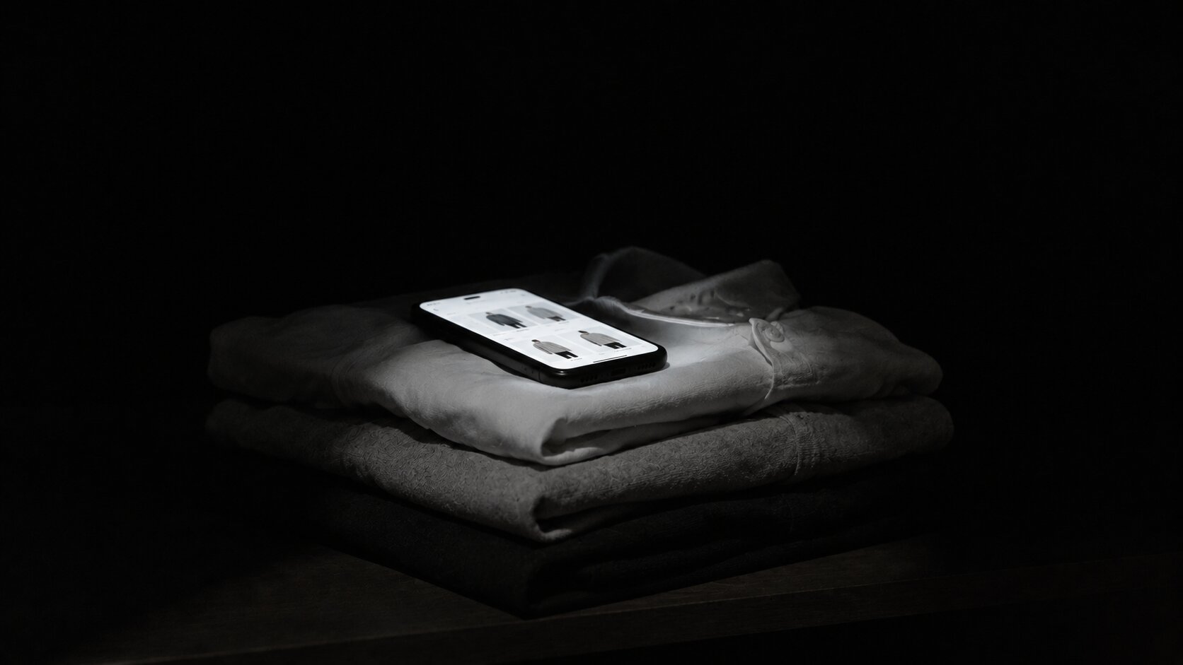 A phone screen glowing on a folded stack of shirts in a dark room, the shirts lit only by the screen.