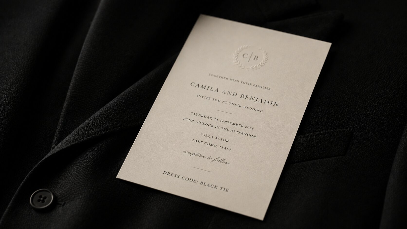 A cream wedding invitation lying face up on a dark tailored jacket, the dress code line at the foot of the card the only text in focus.