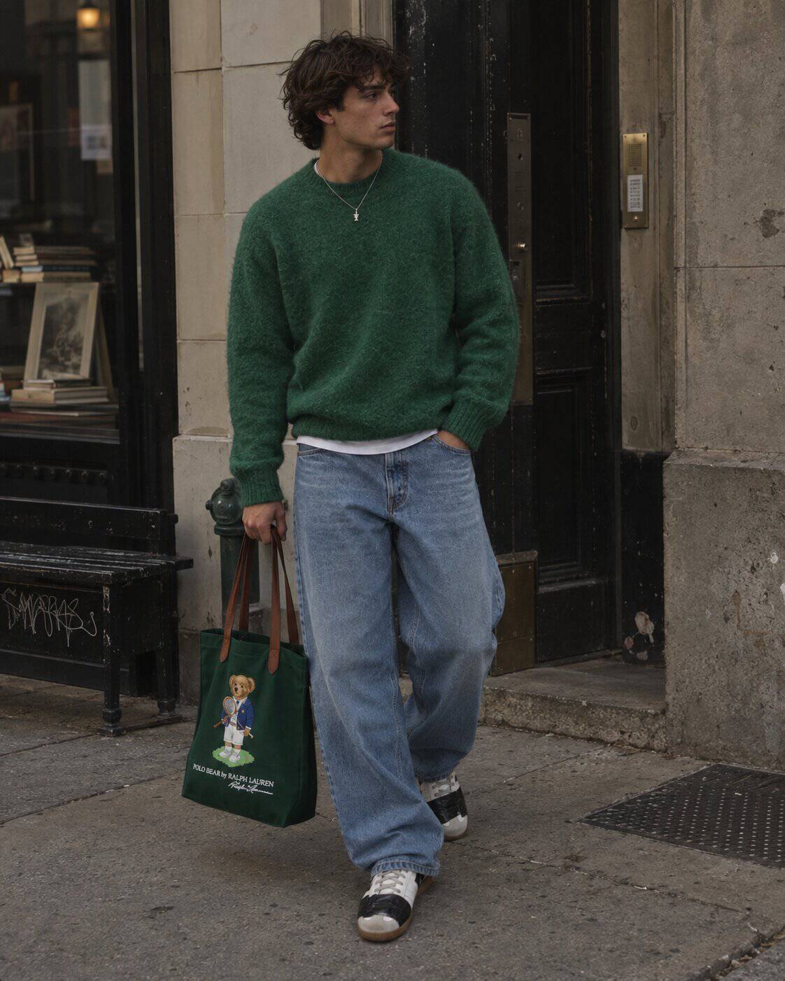 A man in a green brushed Shetland wool sweater over a white T-shirt, wide-leg light-wash distressed jeans and white leather sneakers, carrying a green canvas tote bag on a New York street.