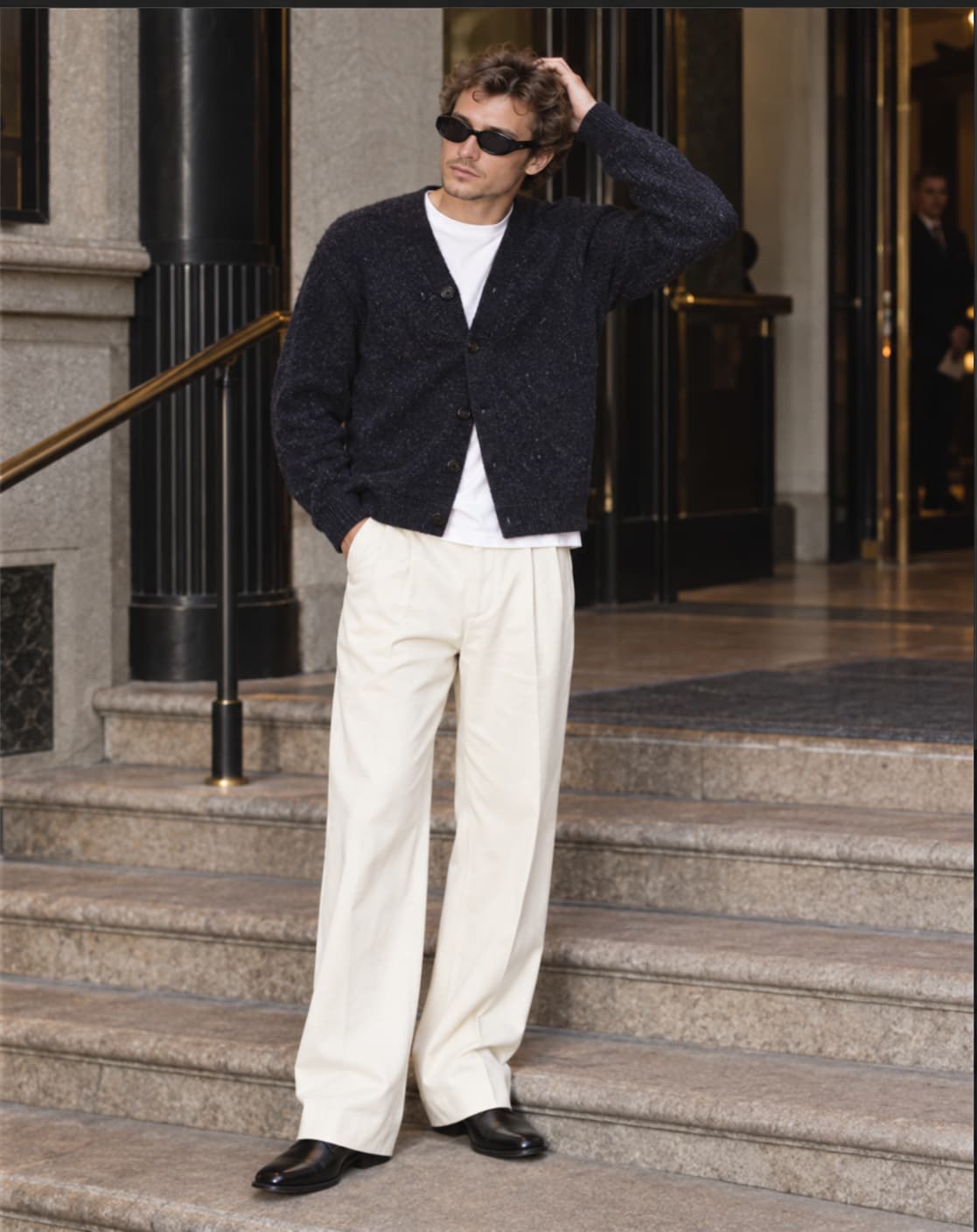 A man in a brushed navy cardigan over a white T-shirt, ecru wide-leg pleated trousers and black square-toed leather boots, wearing black sunglasses on the steps of a hotel in Milan.