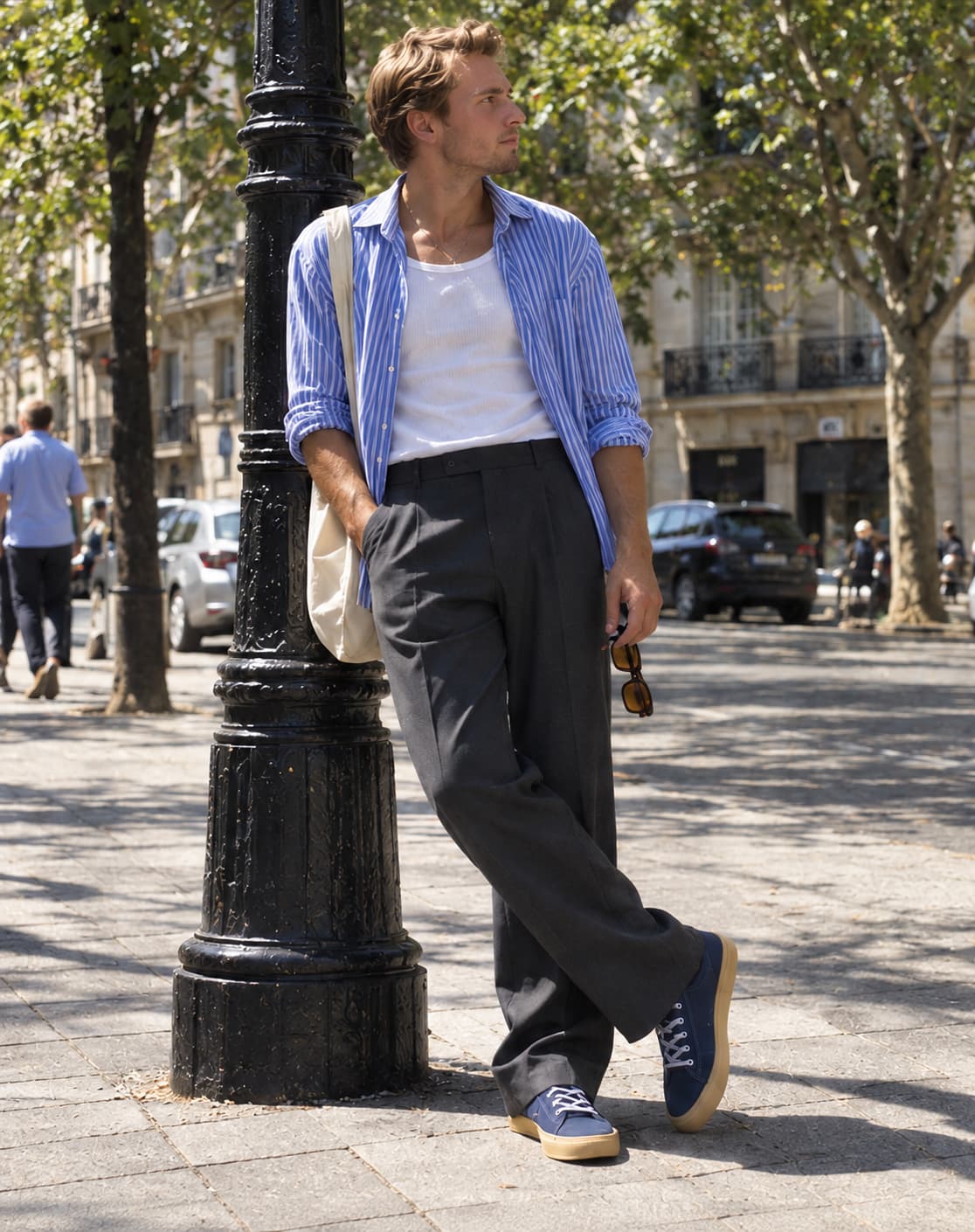 A man in a blue striped cotton shirt worn open over a white ribbed tank top, grey pleated wool trousers and blue plimsolls, holding dark brown sunglasses, leaning against a lamppost on a Barcelona street.